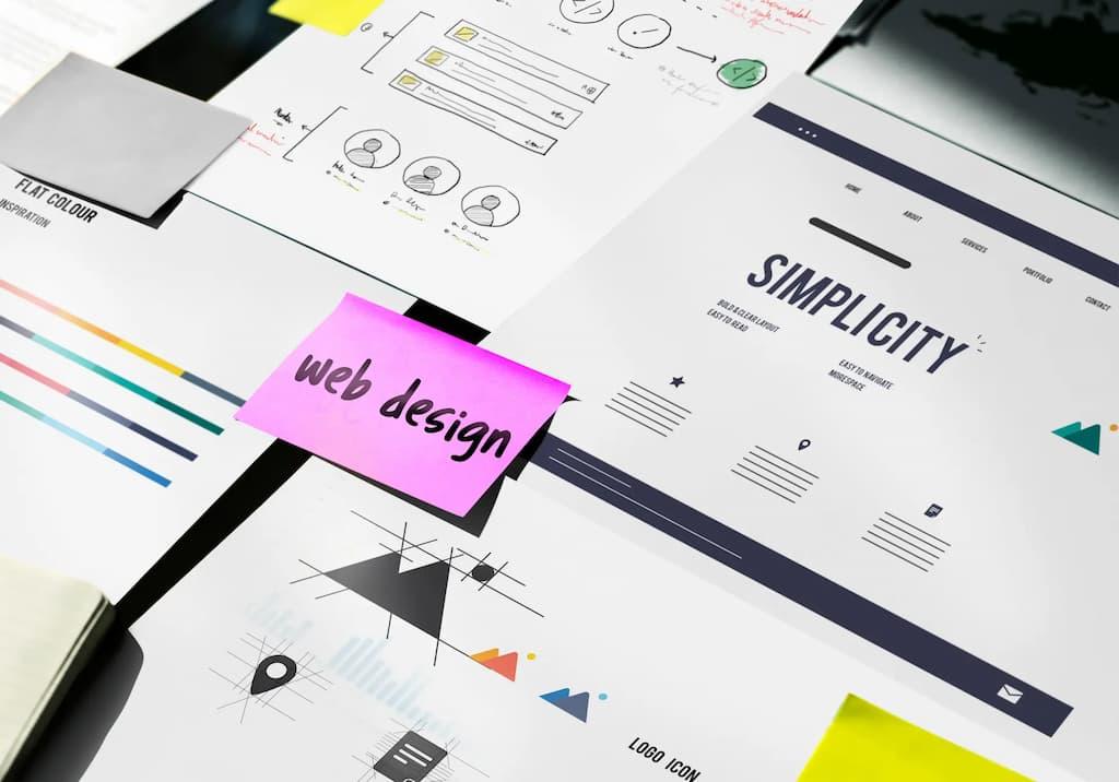 Web design wireframes and sketches on a desk with a pink sticky note and a paper titled Simplicity.